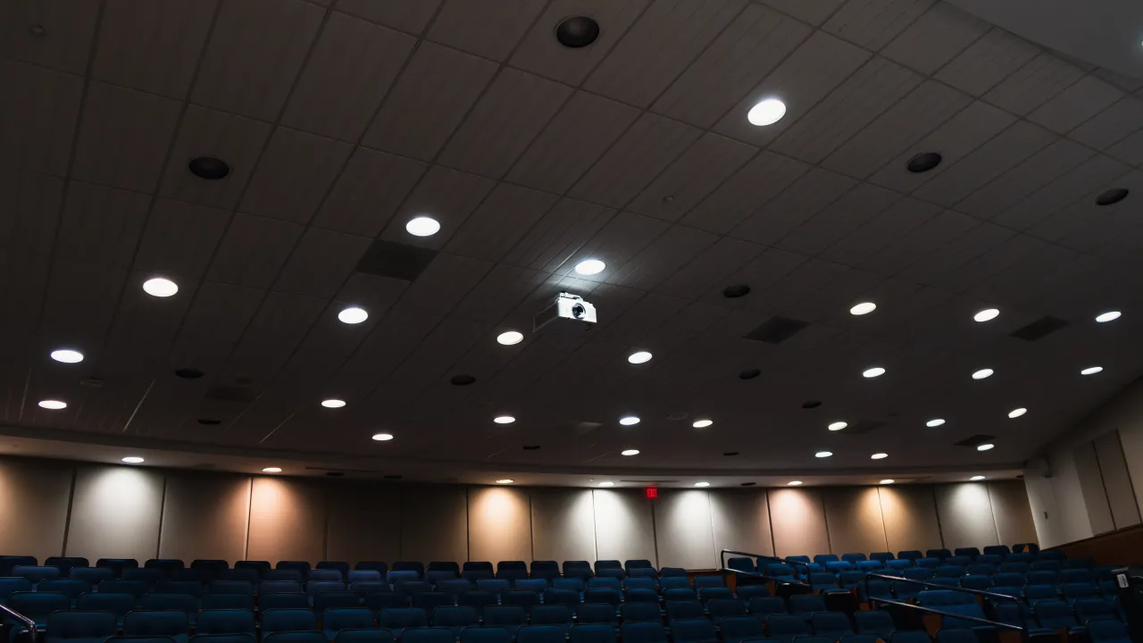 A large-venue laser projector mounted in the ceiling of the College of Business lecture hall, with tiered seating below.