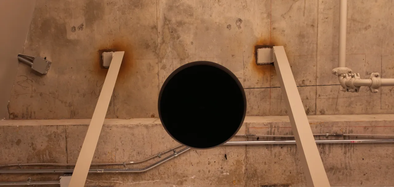 A black pendant loudspeaker seen from below against an exposed-concrete wall.