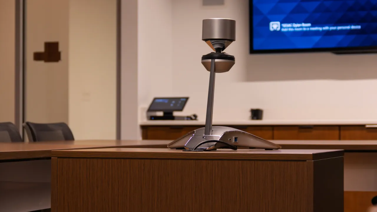A tabletop 360-degree conferencing camera on a meeting-room table, with a wall display behind it.